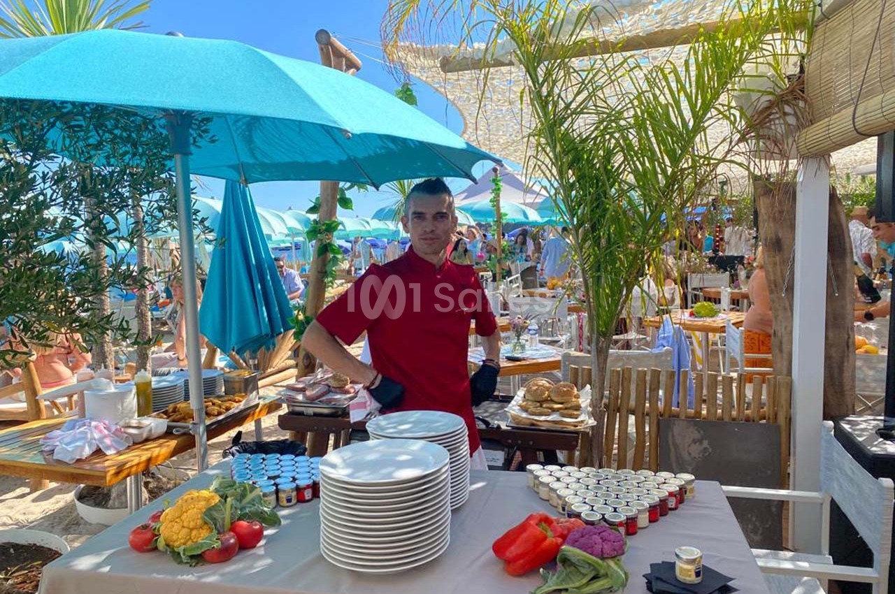 Un cuisinier en tenue rouge devant une table dressée avec des assiettes et des ingrédients, dans un cadre extérieur…