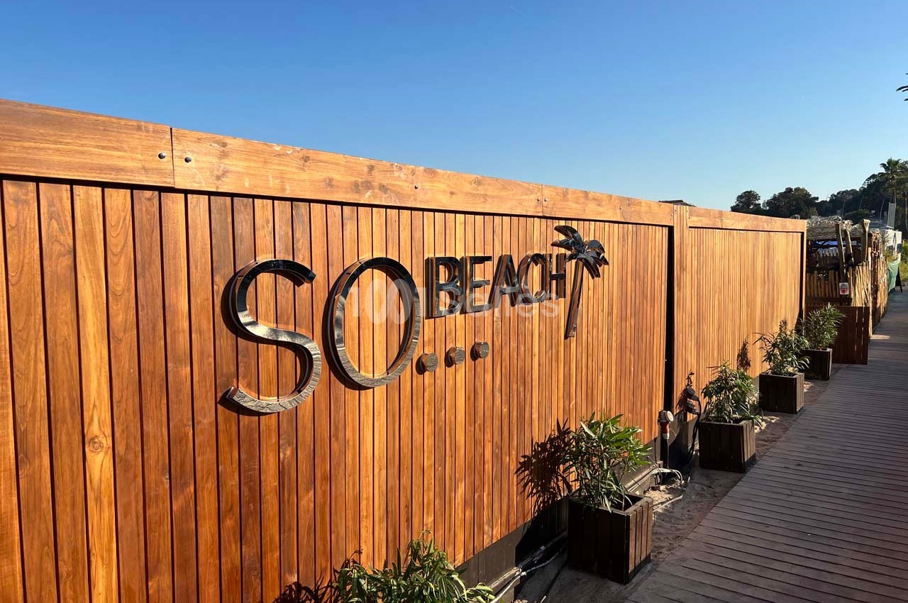 Clôture en bois avec le logo ’SO Beach’ et des plantes en pots alignées le long d'un chemin en bois.