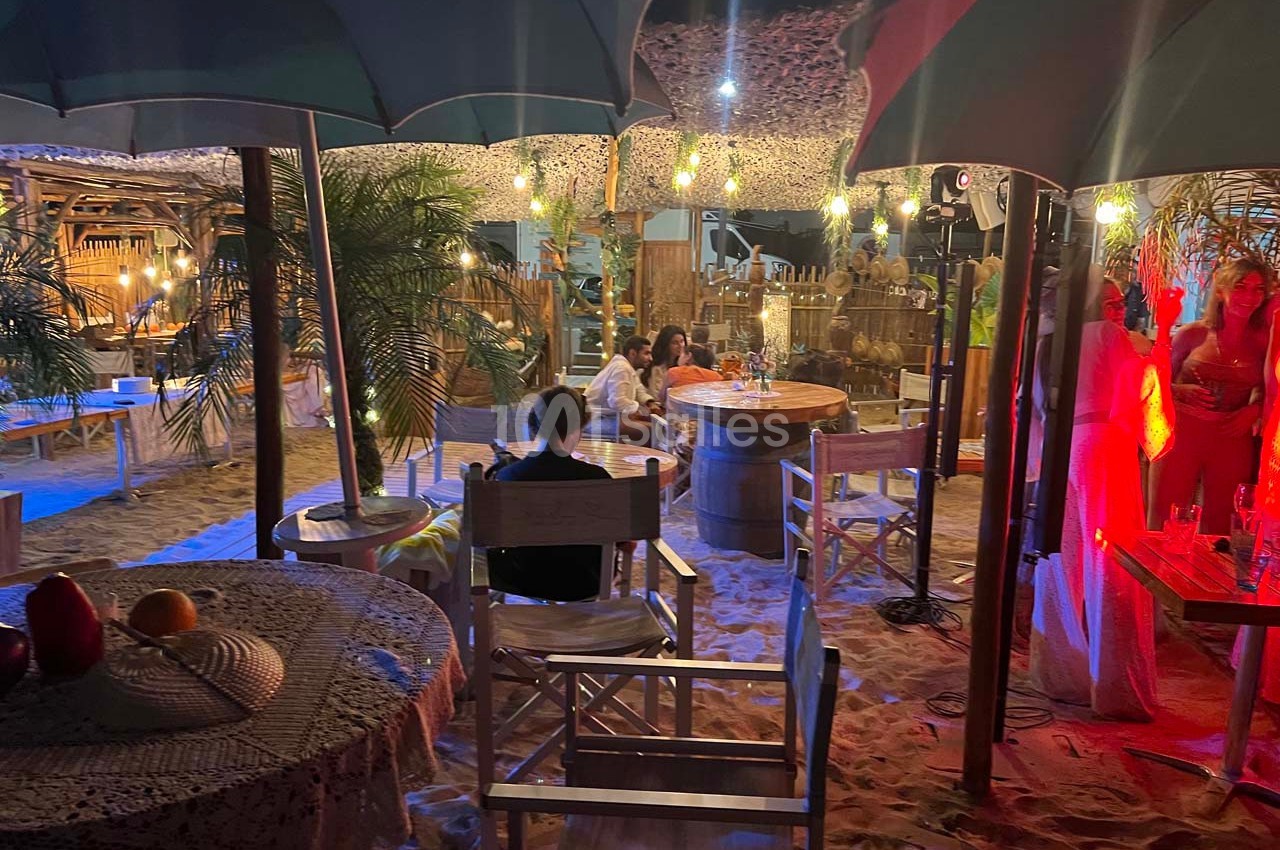 Terrasse de plage éclairée en soirée avec tables, chaises en bois, parasols et ambiance conviviale.