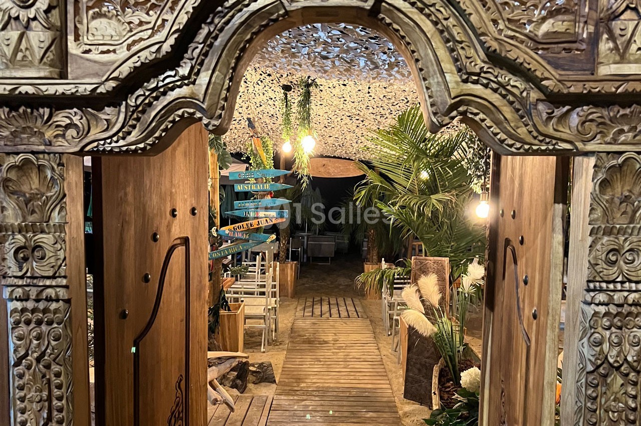 Entrée en bois sculpté menant à un espace extérieur décoré de plantes, lumières suspendues et panneaux directionnels colorés.