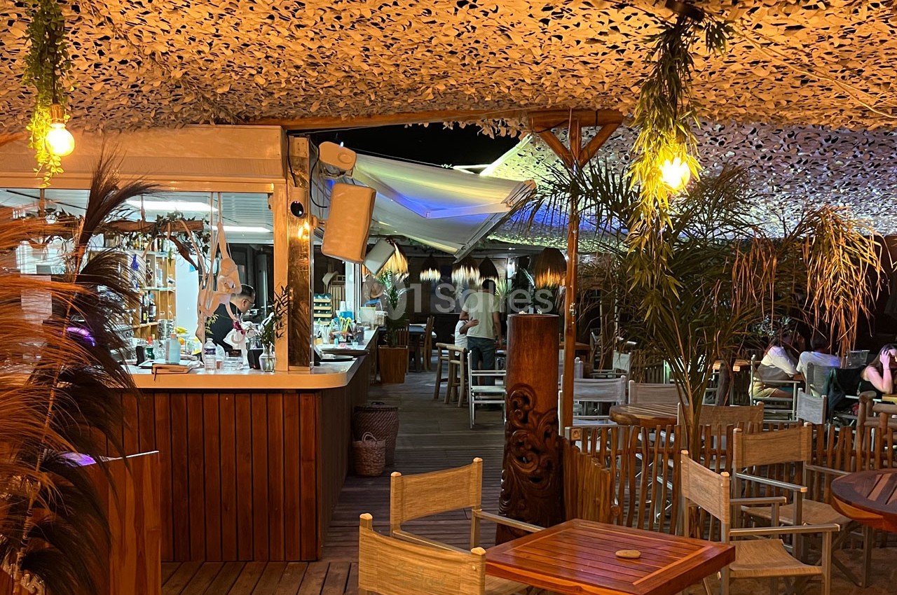 Terrasse extérieure d'un restaurant éclairée en soirée, avec des tables en bois et un bar en arrière-plan.