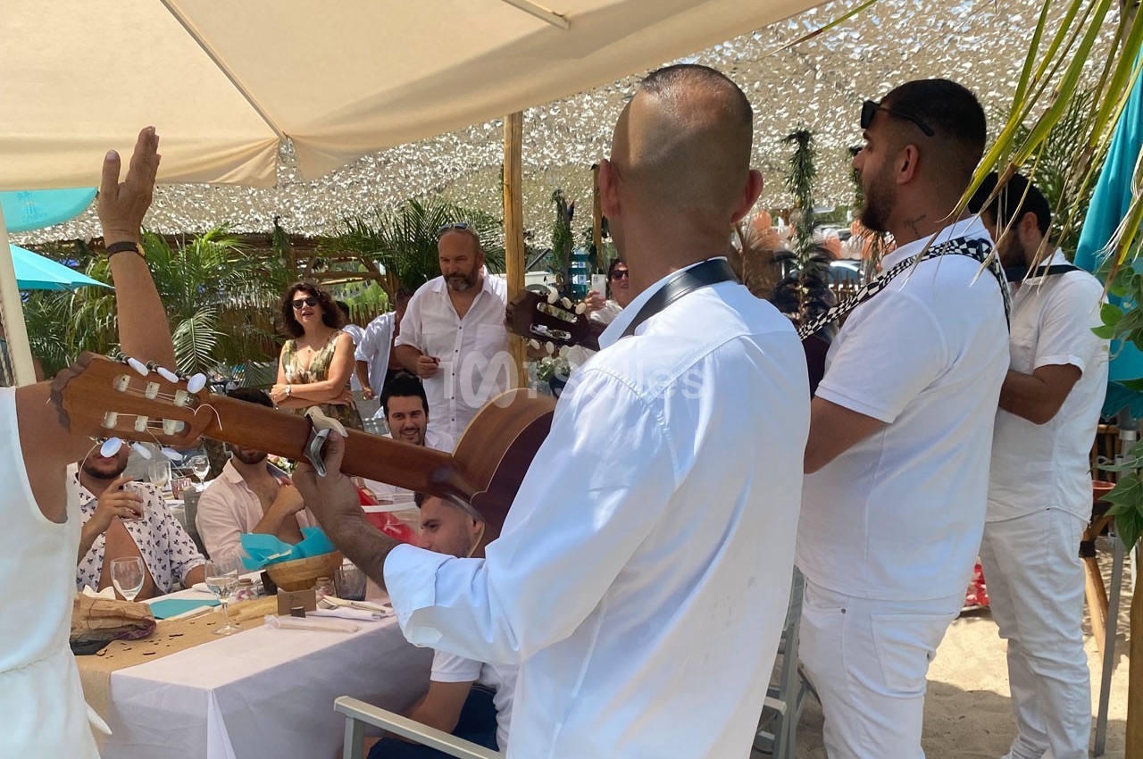 Musiciens jouant de la guitare et animant un repas en plein air sous des parasols, entourés de convives.