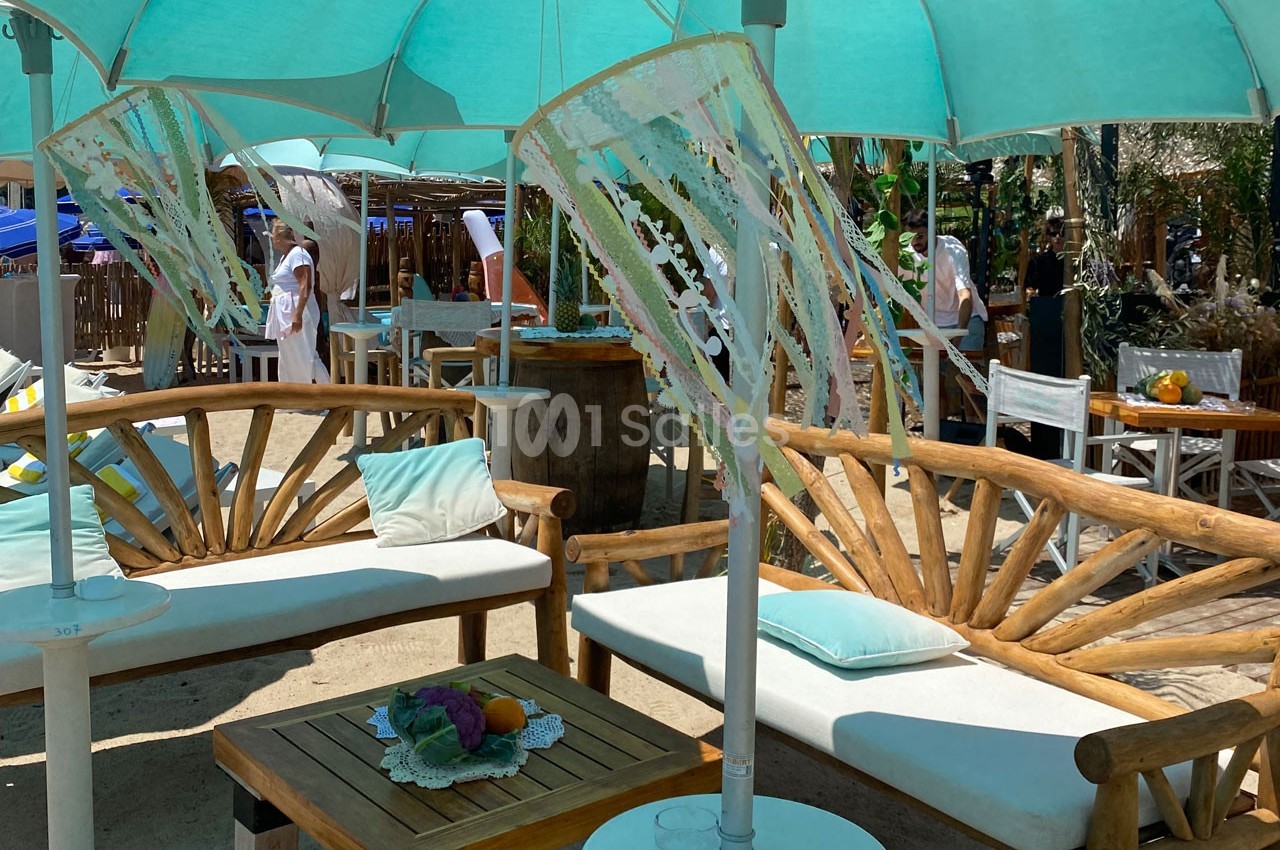 Mobilier en bois avec coussins sous des parasols turquoise sur une terrasse ensoleillée près d'une plage.