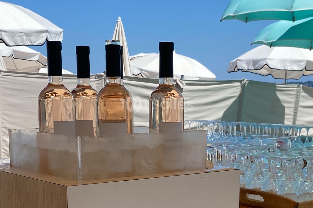 Bouteilles de vin rosé sur glace devant des parasols blancs et bleus sous un ciel dégagé.
