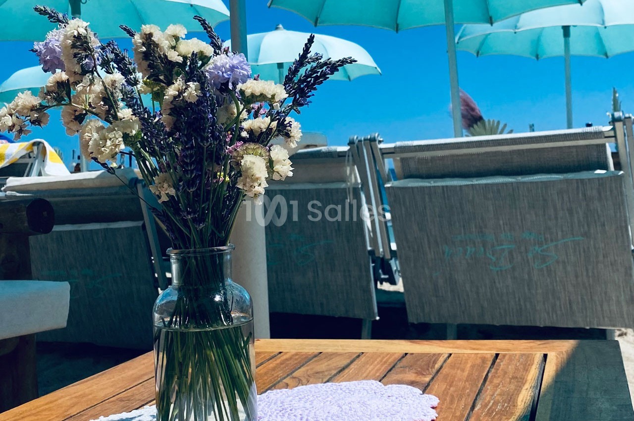 Vase de fleurs sauvages sur une table en bois, avec des parasols bleus et des transats en arrière-plan sur une plage.