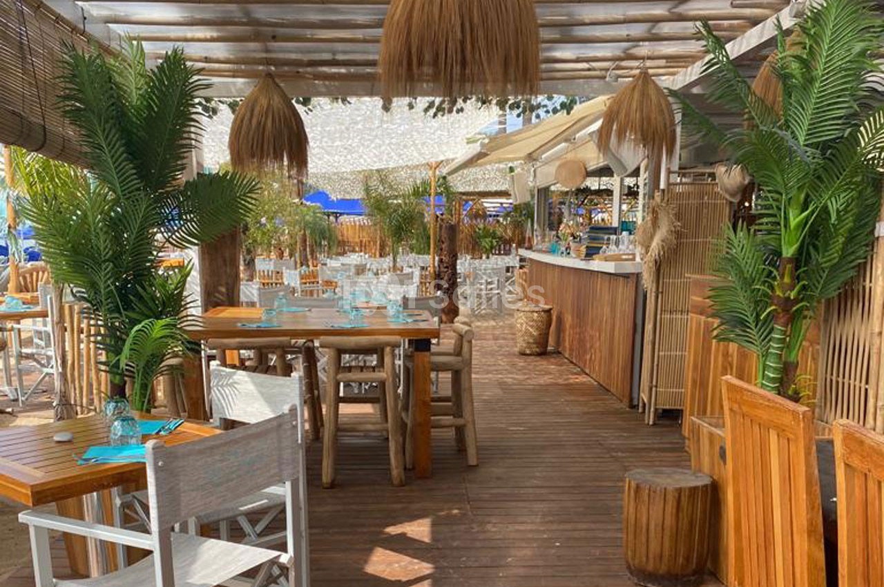 Terrasse en bois d'un restaurant avec tables, chaises, plantes tropicales et luminaires suspendus en paille.