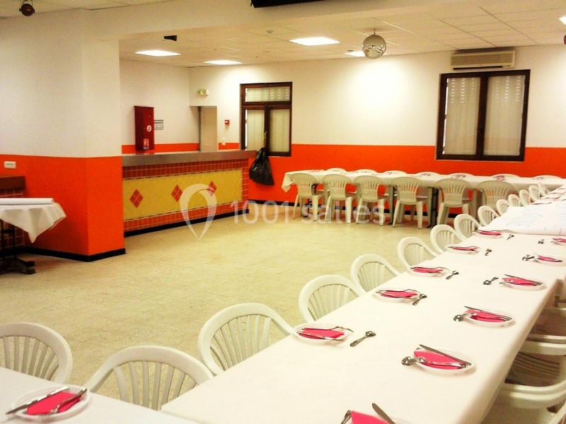 Salle lumineuse avec tables alignées, nappes blanches, chaises en plastique, et un comptoir en fond.