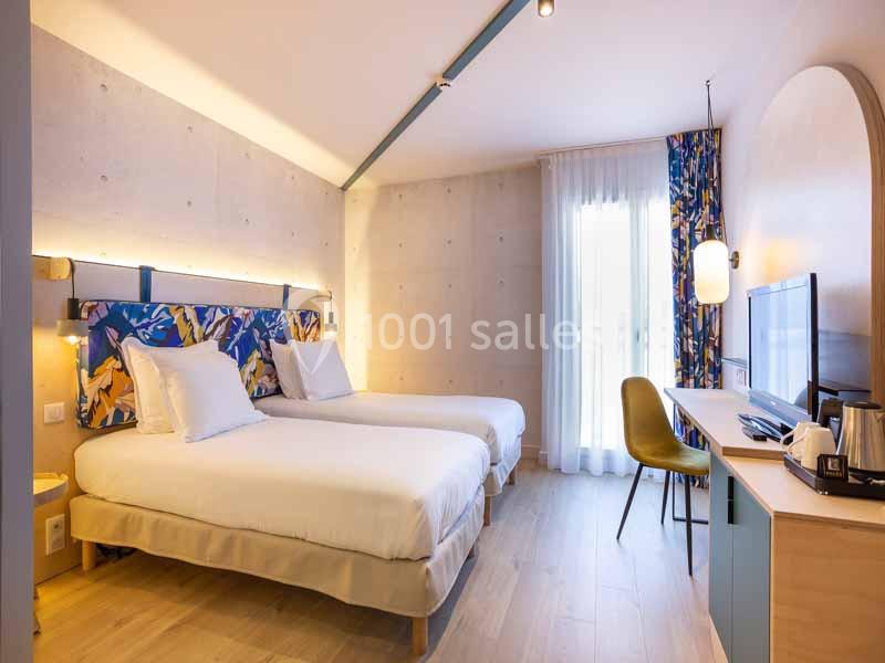 Chambre lumineuse avec deux lits simples, bureau, télévision, chaise verte et grande fenêtre avec rideaux colorés.