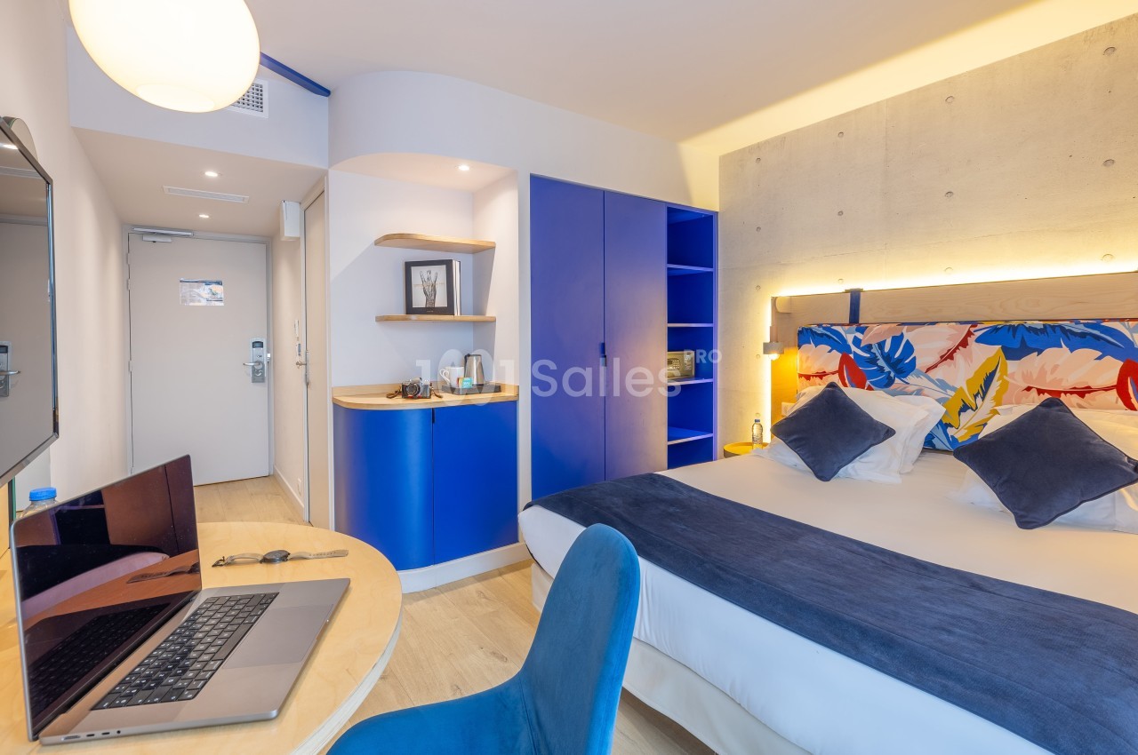 Chambre d'hôtel moderne avec lit double, bureau équipé d'un ordinateur portable et rangements aux tons bleus.