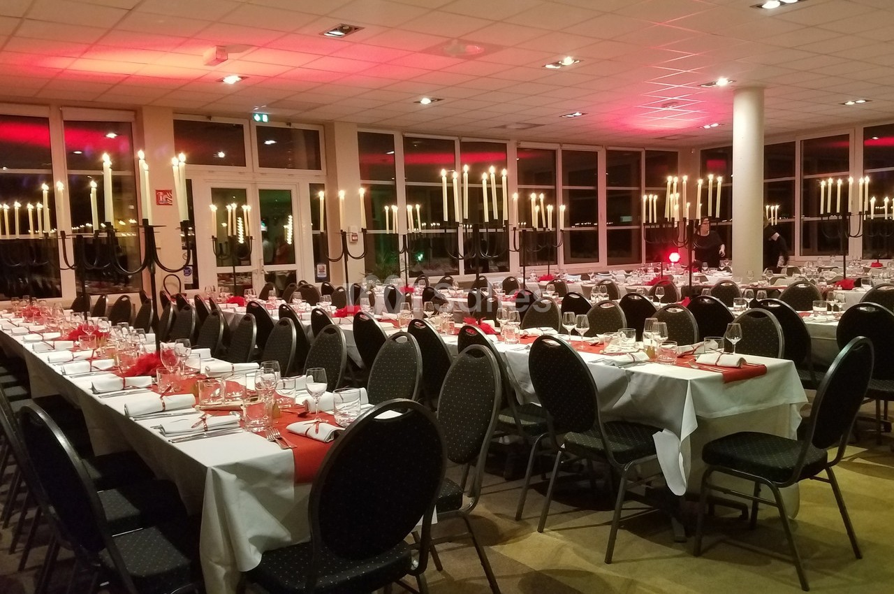 Salle de réception décorée avec des chandeliers, des tables dressées avec nappes blanches et serviettes rouges.