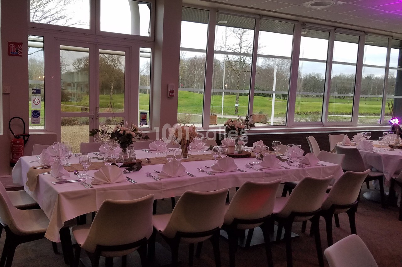 Salle de réception avec tables dressées, nappes blanches, décorations florales et vue sur un extérieur verdoyant.