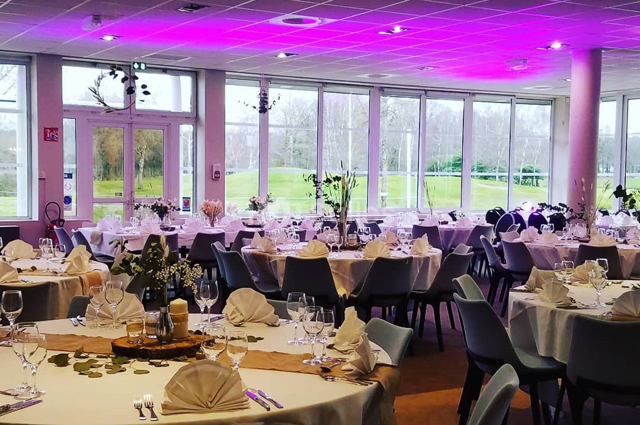 Salle de réception décorée pour un événement avec tables dressées, nappes blanches et éclairage rose au plafond.