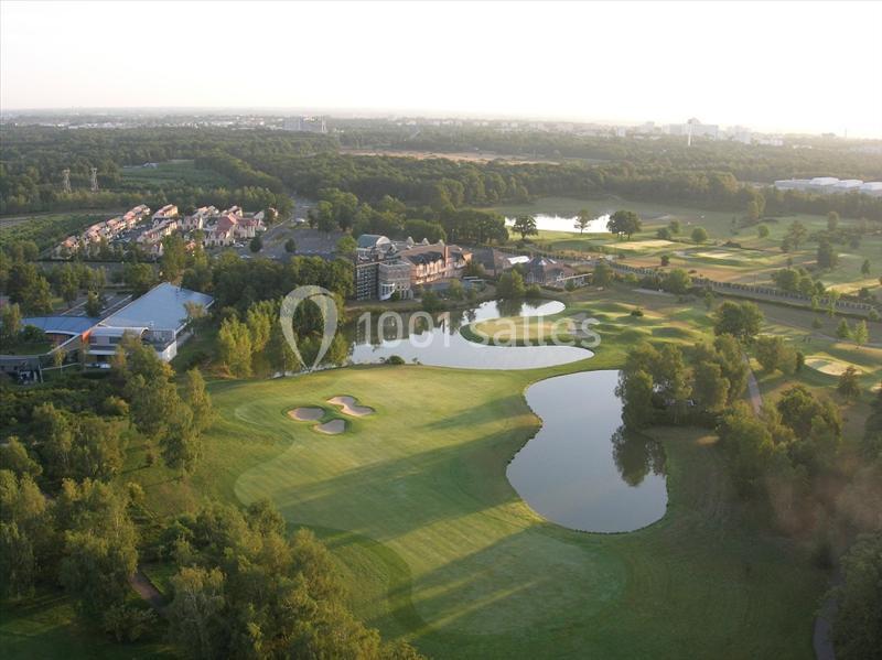 Vue aérienne d'un terrain de golf avec des plans d'eau, des bunkers, des arbres et des bâtiments en arrière-plan.
