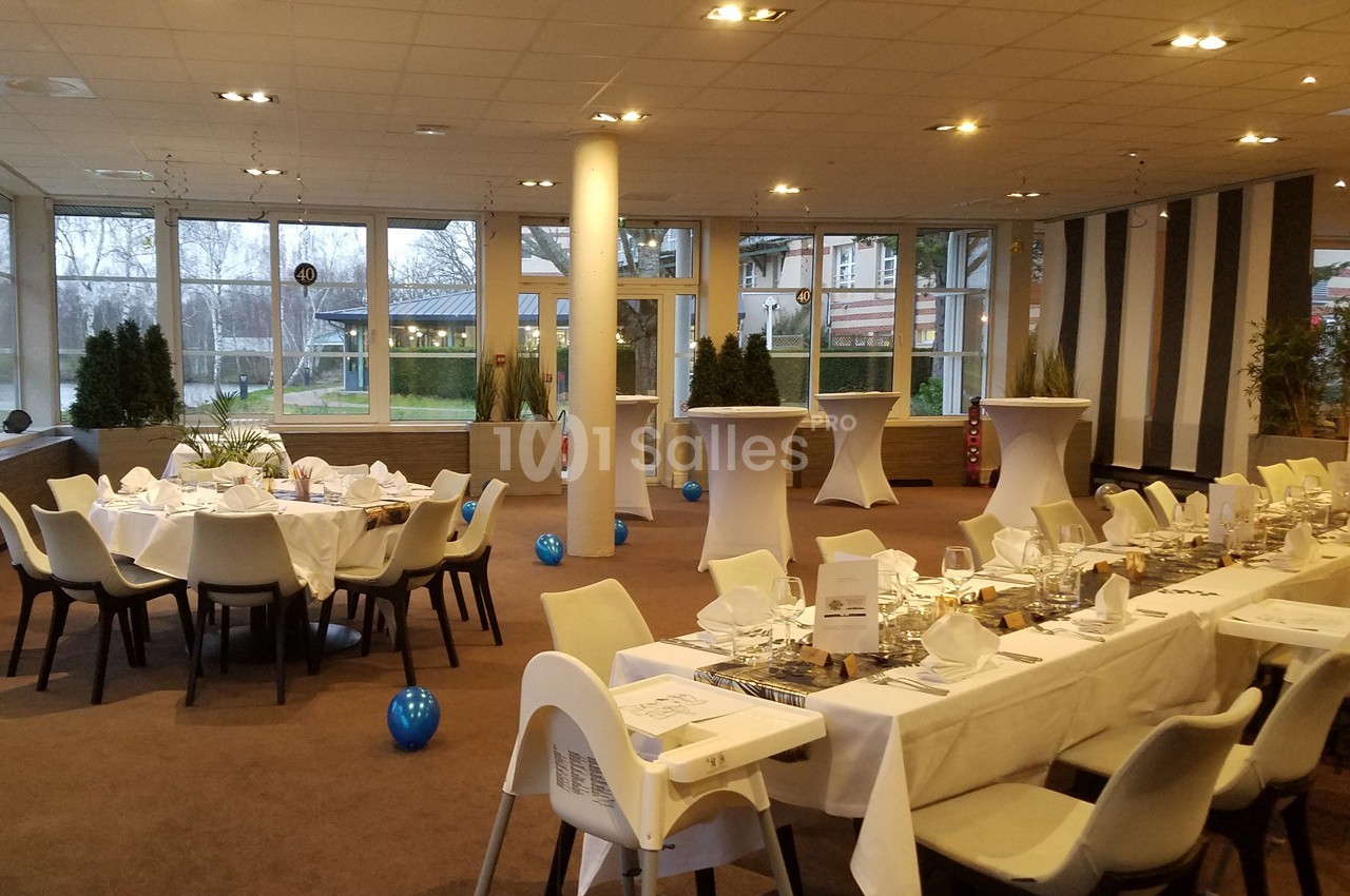 Salle de réception lumineuse avec tables dressées pour un repas, décorée de ballons bleus et entourée de grandes baies…