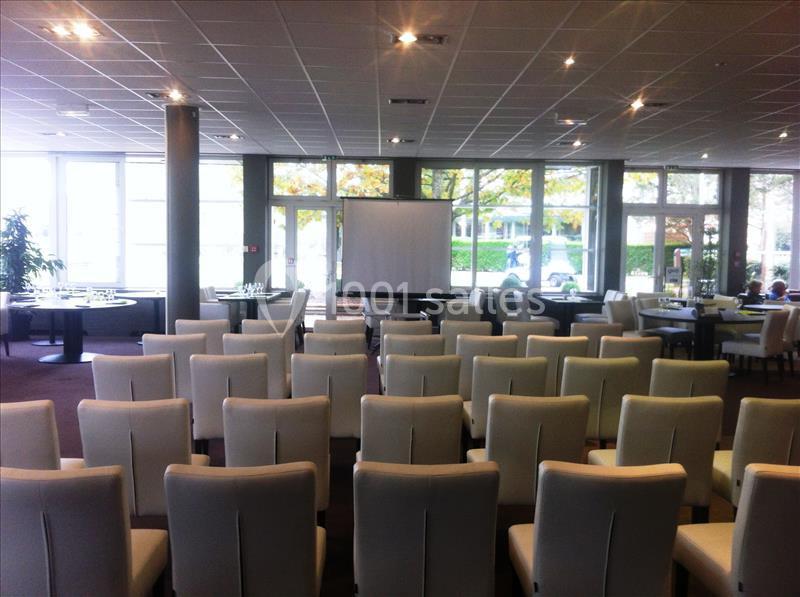 Salle de conférence avec des rangées de chaises blanches alignées face à un écran de projection.