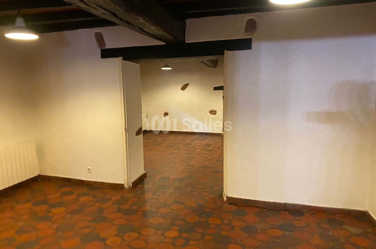 Salle vide avec carrelage en terre cuite, murs blancs, poutres apparentes au plafond et éclairage suspendu.