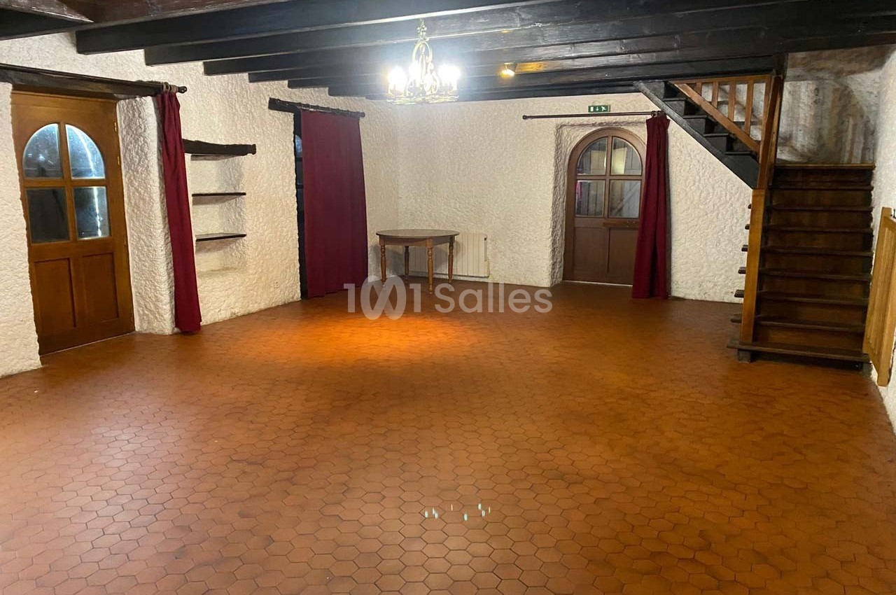Grande pièce vide avec sol en carrelage hexagonal, murs clairs, poutres apparentes, escaliers en bois et rideaux rouges.