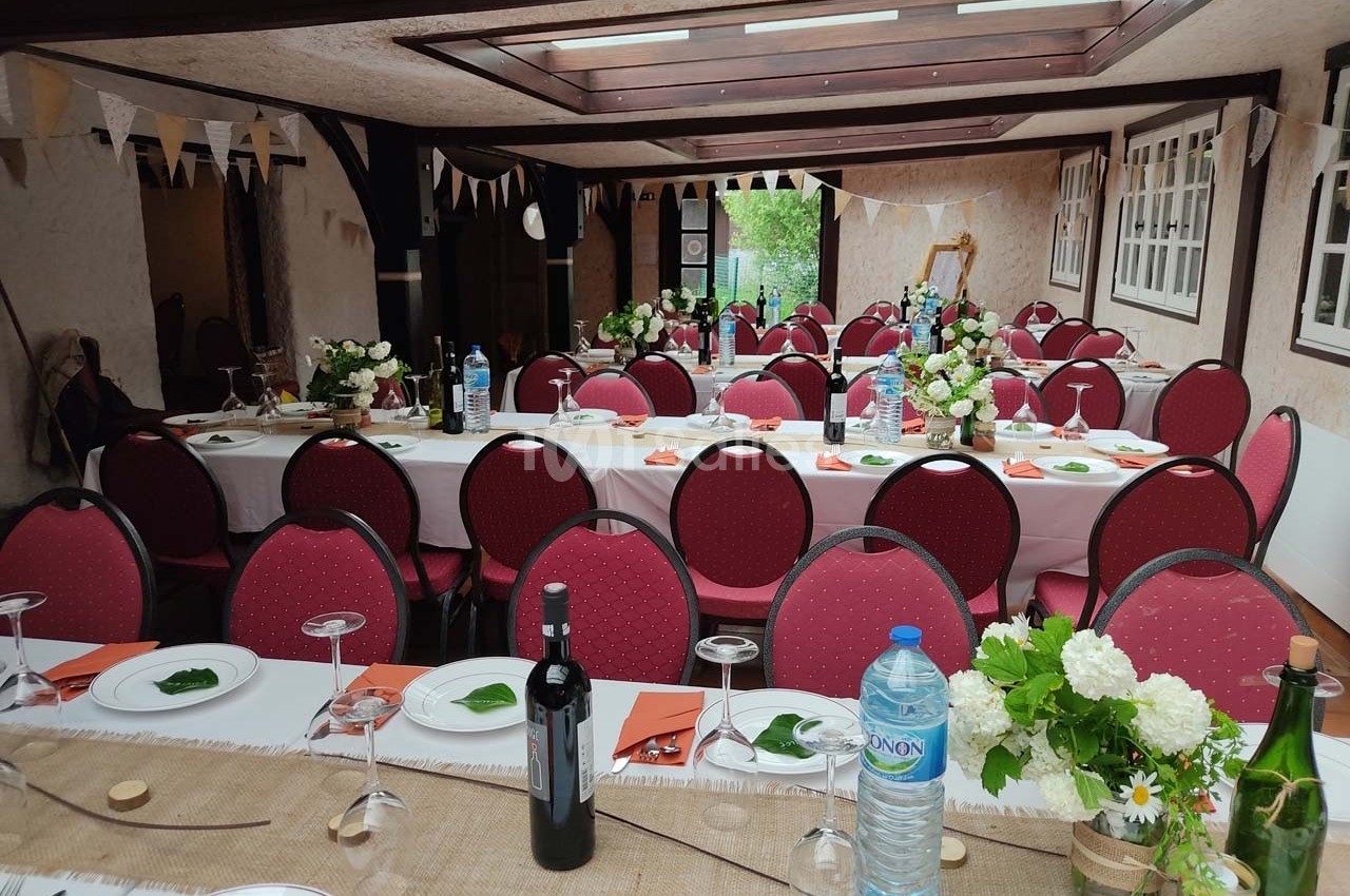 Salle décorée pour un repas festif avec tables dressées, chaises rouges, fleurs, bouteilles et guirlandes suspendues.