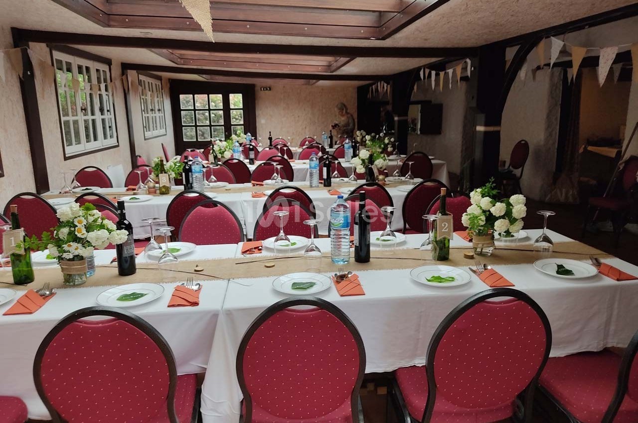 Salle décorée pour un événement avec des tables alignées, nappes blanches, chaises rouges et centres de table fleuris.