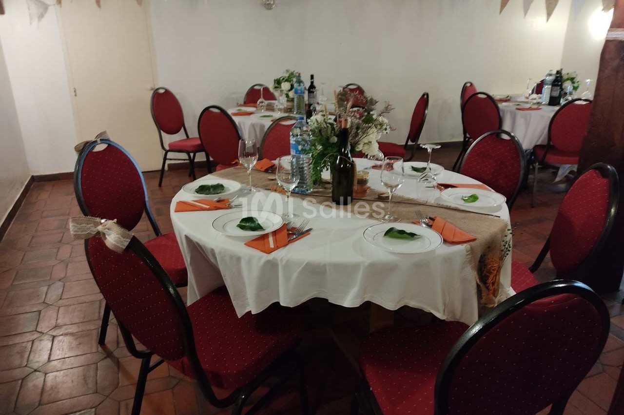 Salle décorée pour un repas, avec des tables rondes dressées, nappes blanches et chaises rouges.