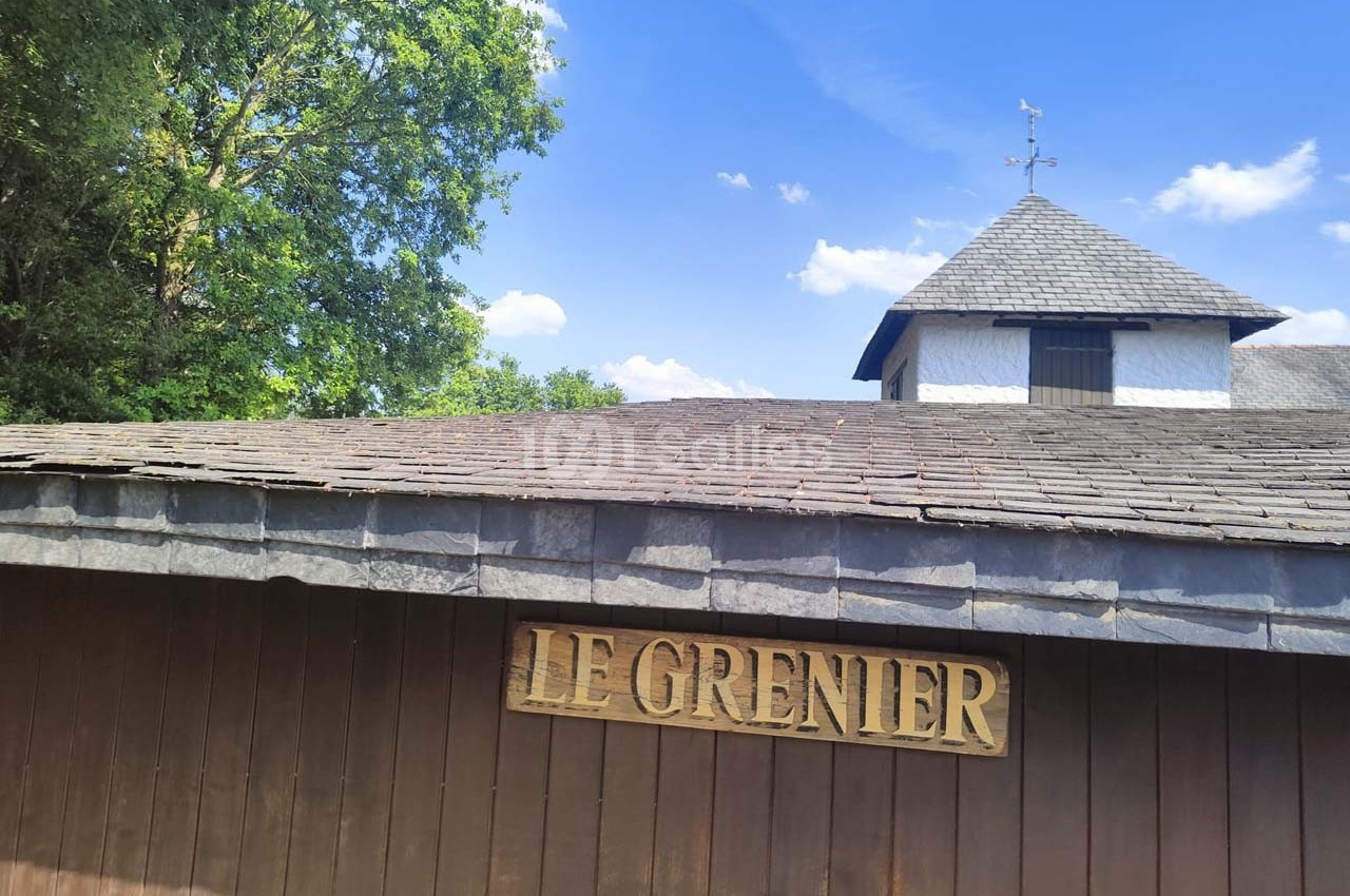 Façade en bois avec une enseigne ’Le Grenier’, sous un toit en ardoise, entourée de végétation et ciel bleu.