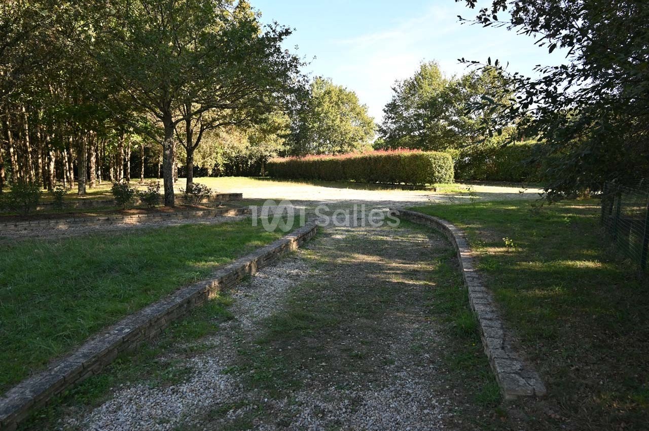 Allée en gravier bordée de pelouses et d'arbres, menant à une haie et une clairière ensoleillée.