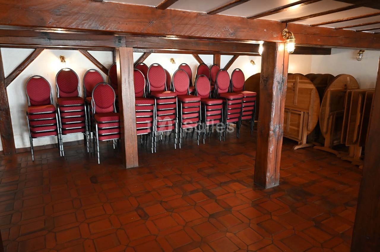 Chaises empilées et tables pliées rangées dans une salle au sol en carrelage et poutres apparentes.