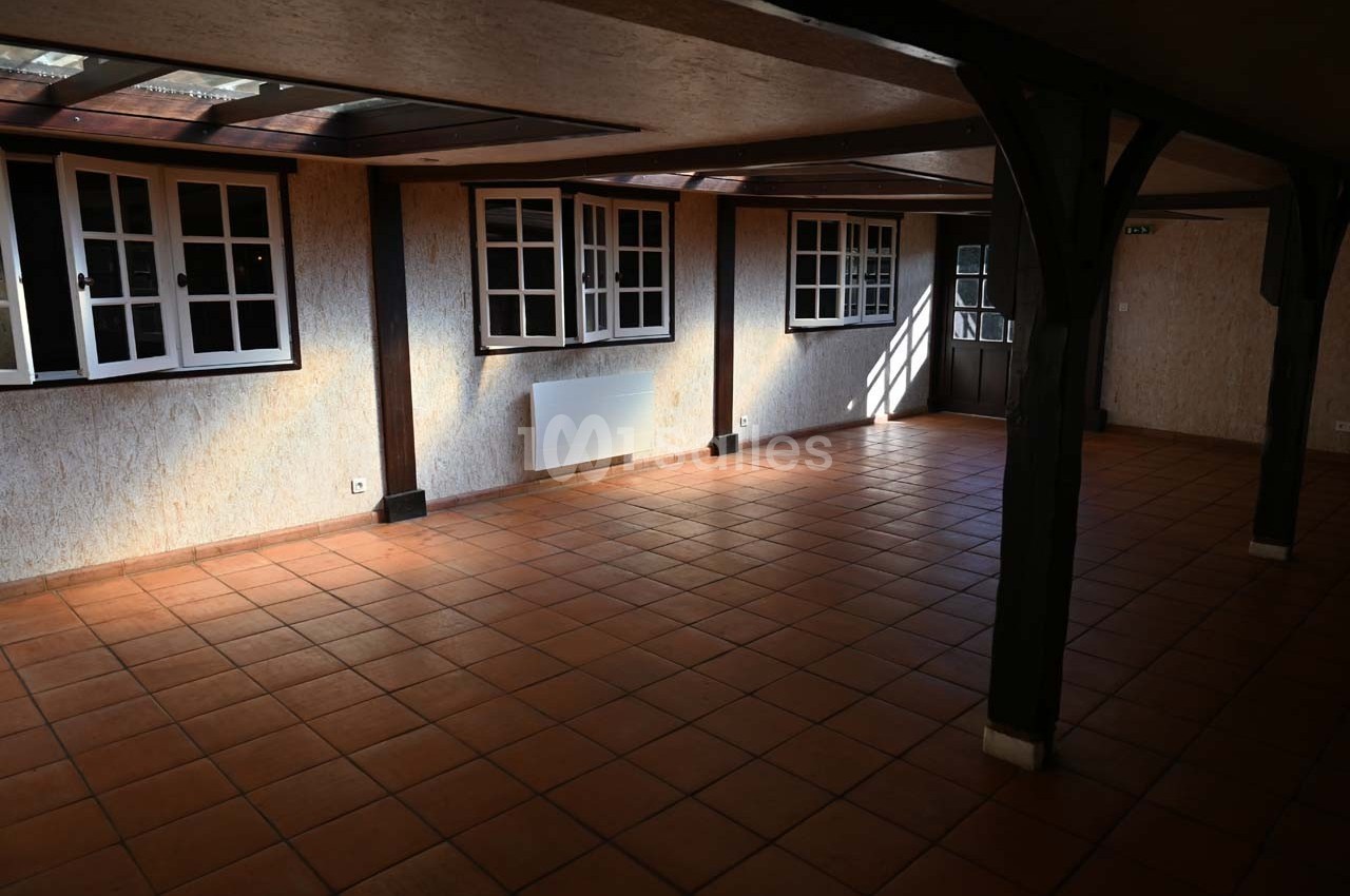 Salle vide avec carrelage en terre cuite, murs clairs, fenêtres à carreaux et poutres en bois apparentes.