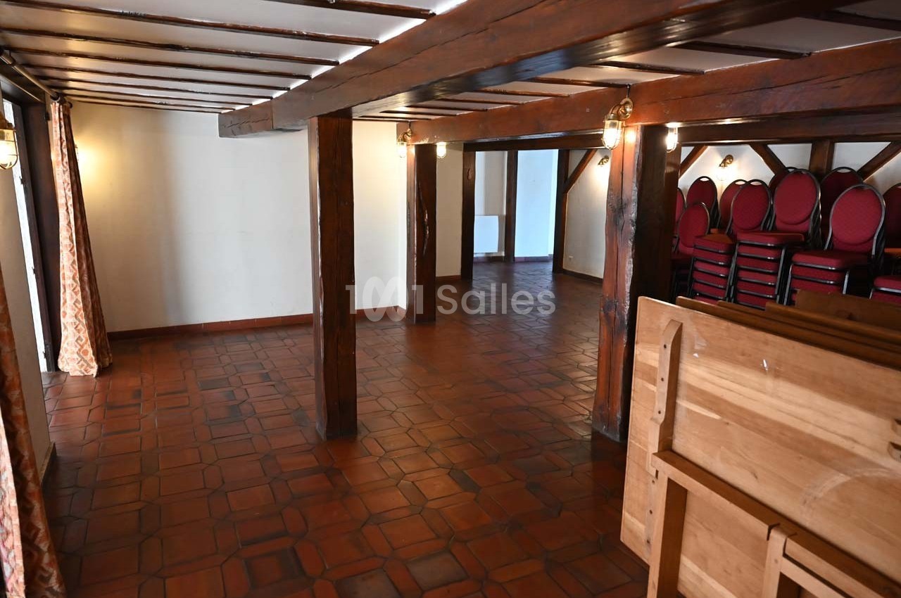 Salle vide avec sol en carrelage, poutres apparentes, chaises empilées dans un coin et lumière naturelle entrant par les…