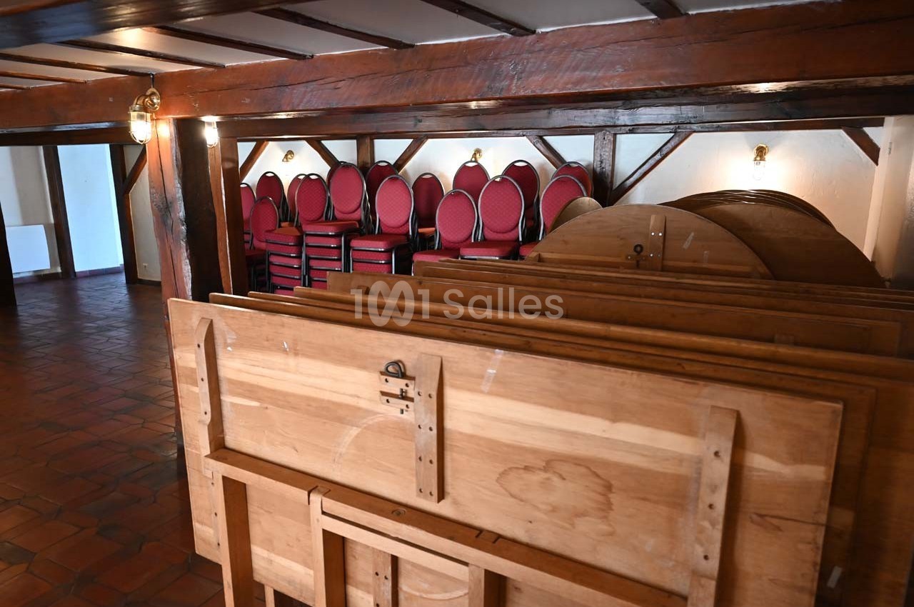 Tables pliées en bois empilées au premier plan, avec des chaises rouges empilées contre un mur en arrière-plan.