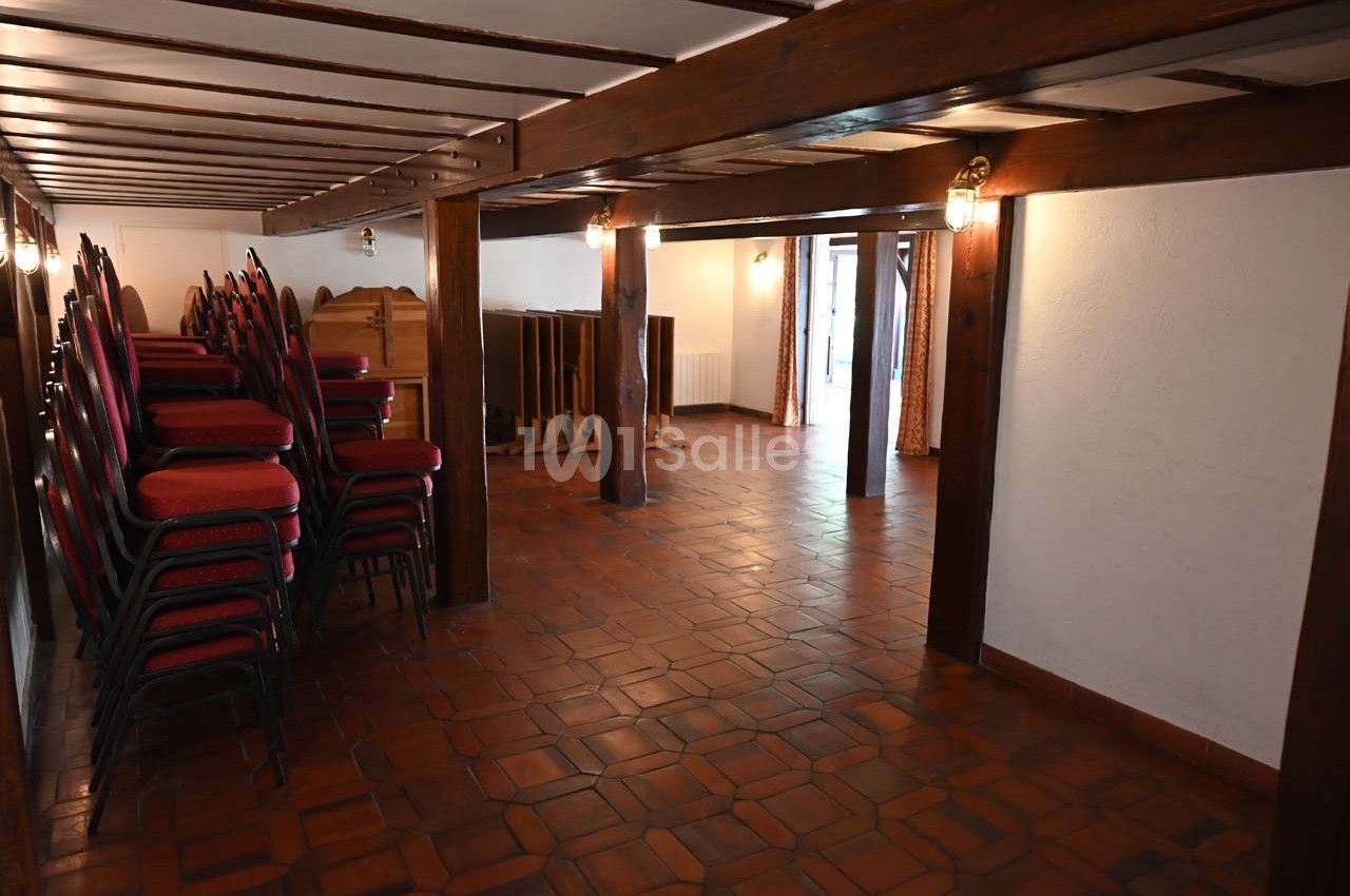 Salle vide avec sol en carrelage, poutres apparentes, chaises empilées et lumière naturelle entrant par une porte vitrée.