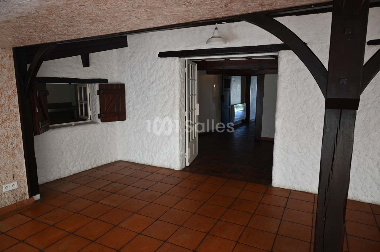Salle vide avec murs blancs texturés, poutres en bois sombre, carrelage au sol et une ouverture vers une autre pièce.