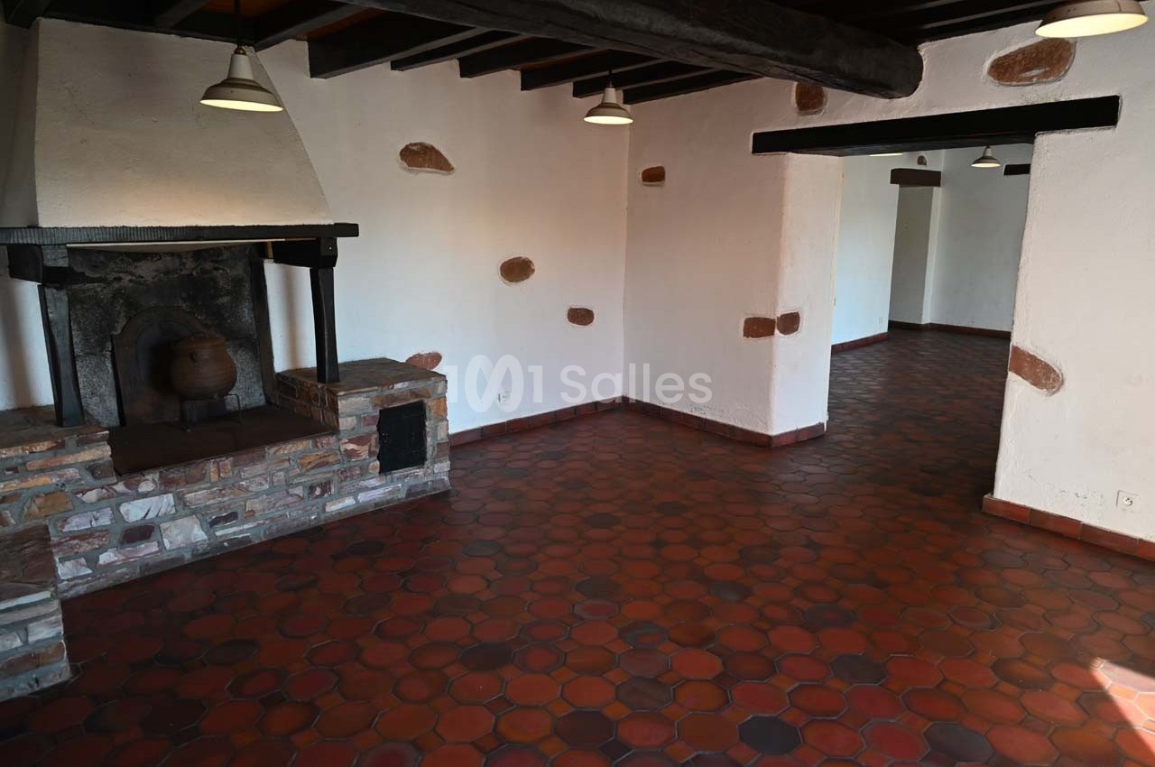 Intérieur d'une pièce vide avec sol en tomettes rouges, cheminée en pierre et poutres apparentes au plafond.