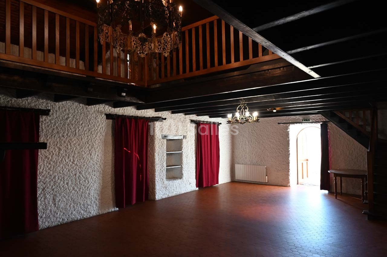 Salle intérieure avec murs en pierre blanche, rideaux rouges, poutres apparentes et lustres suspendus.