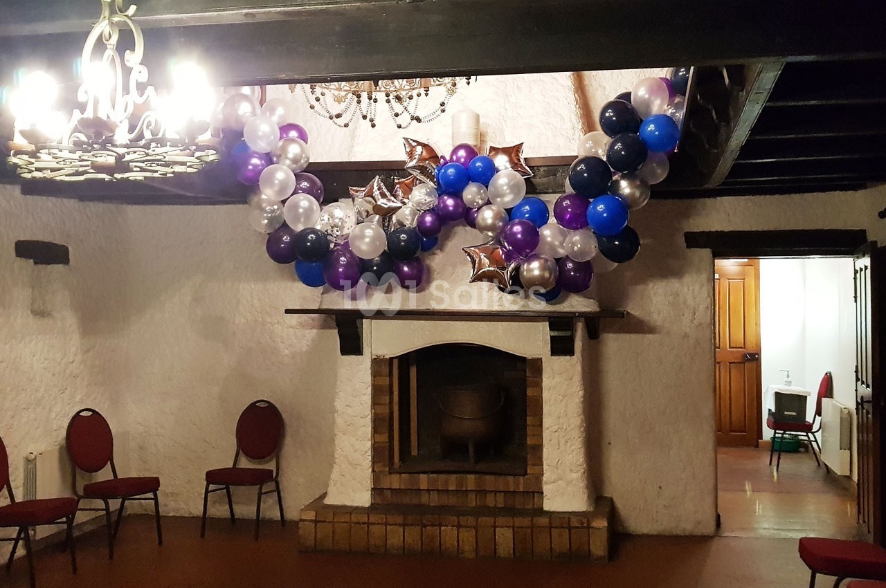 Cheminée décorée avec des ballons colorés et des étoiles argentées dans une salle avec des chaises et un lustre.