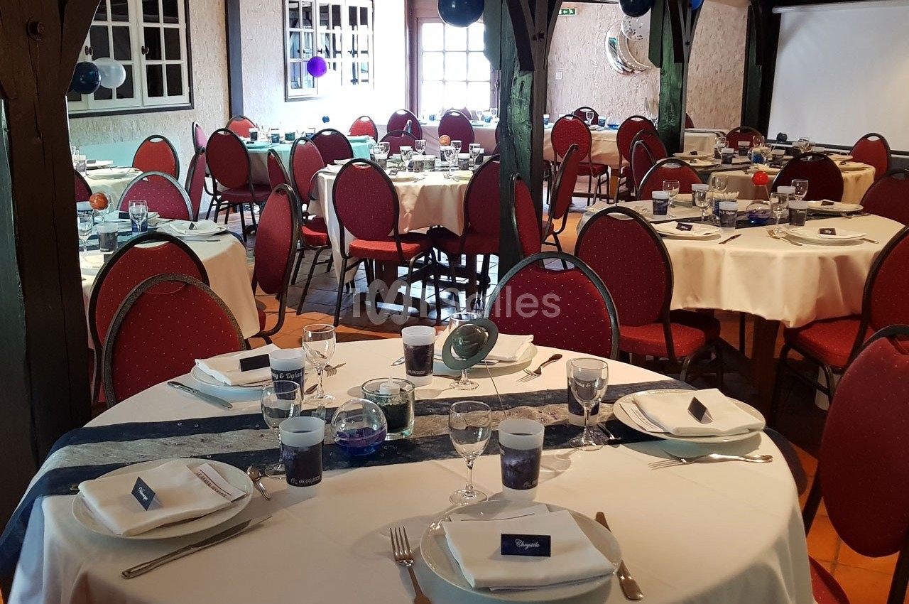 Salle de réception avec tables rondes dressées, nappes blanches, chaises rouges et décorations festives.