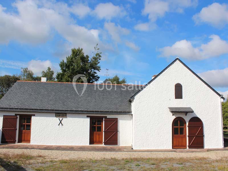 Bâtiment blanc à toit d'ardoise avec portes en bois, entouré de végétation sous un ciel bleu partiellement nuageux.