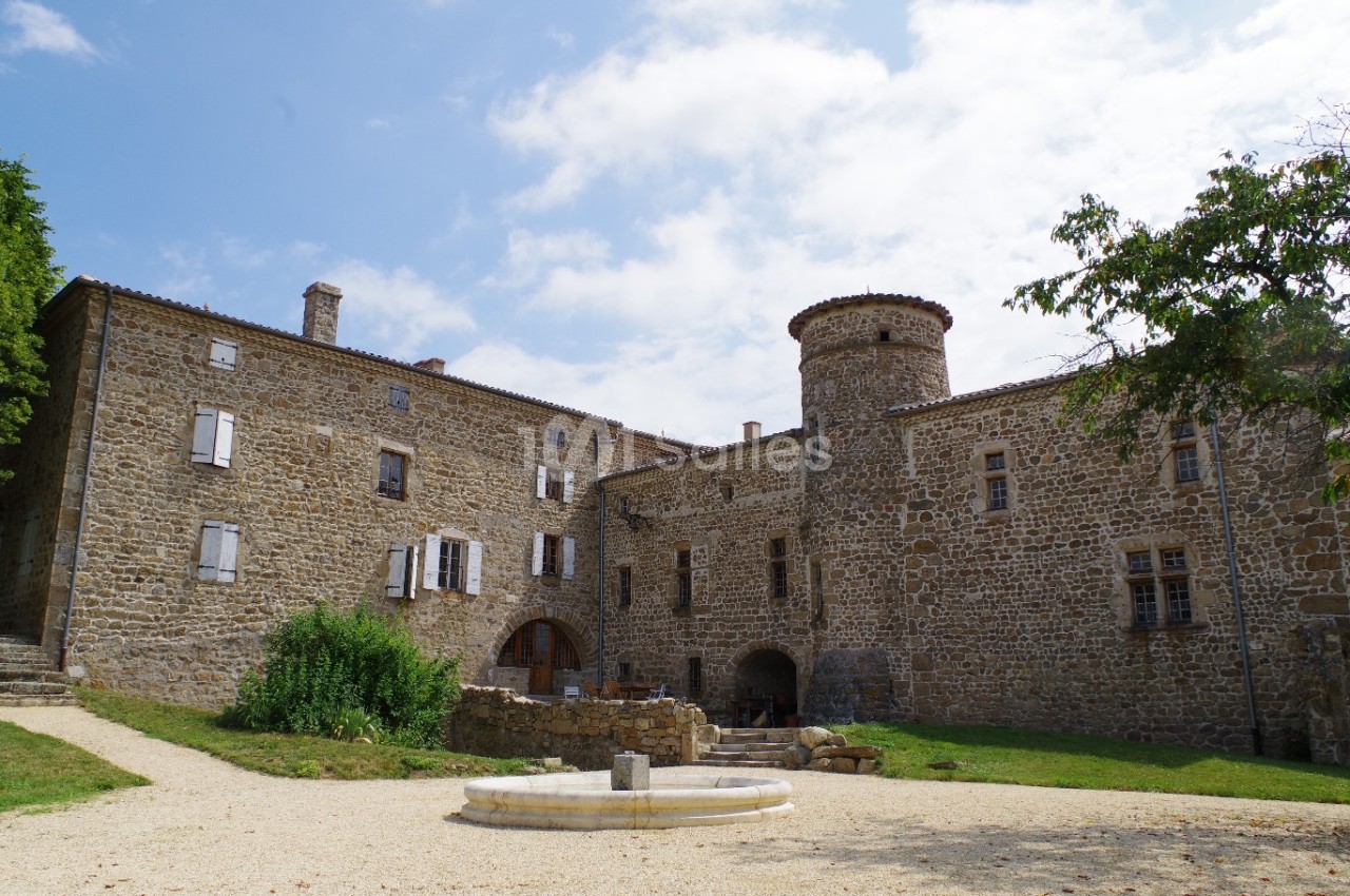Façade en pierre d'un château historique avec une tour ronde, entouré de verdure et d'une cour avec une fontaine.