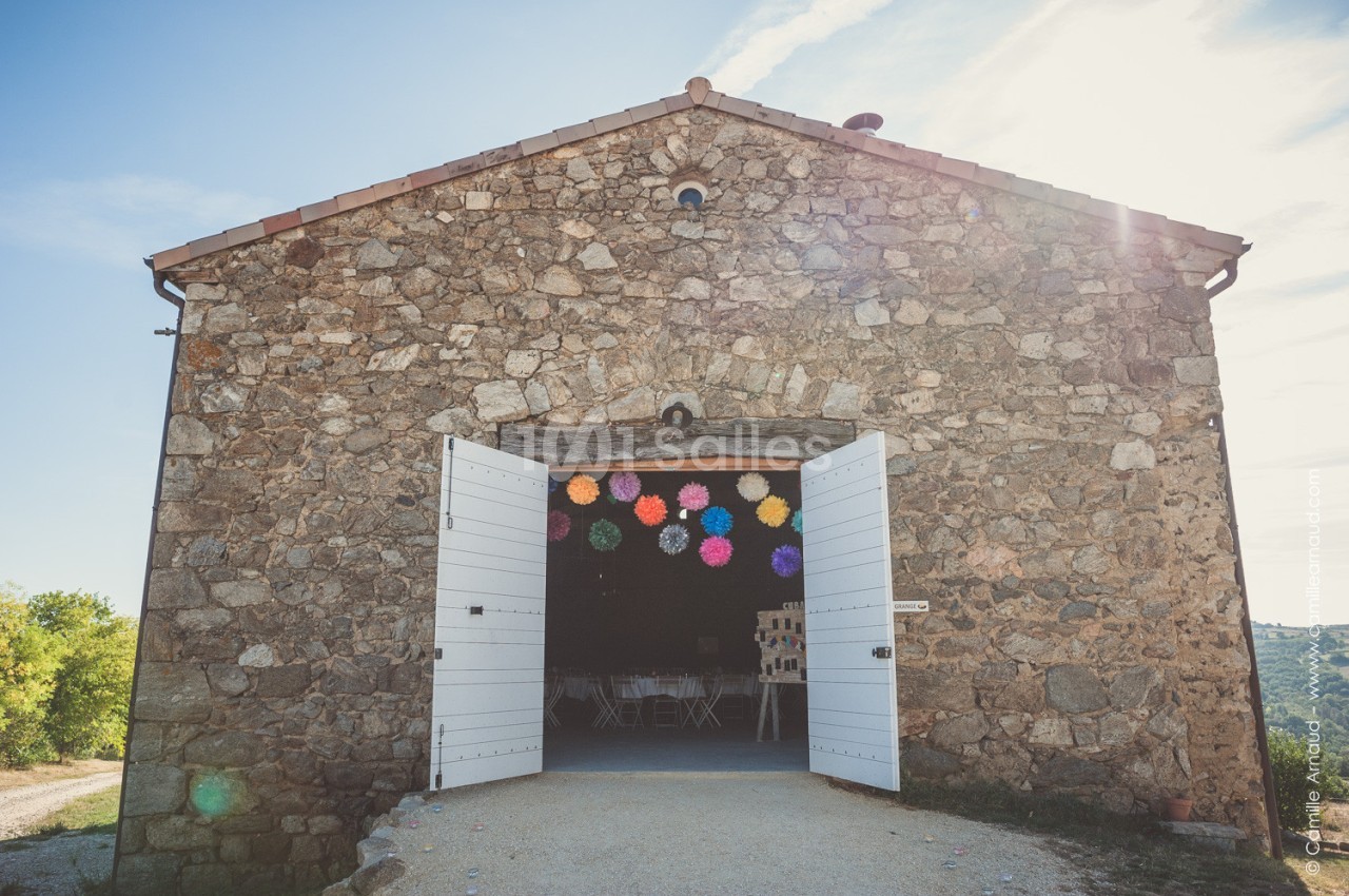 Façade en pierre d'une grange avec portes blanches ouvertes, décorée de pompons colorés suspendus à l'intérieur.