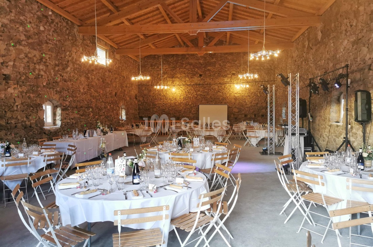Salle en pierre avec plafond en bois, tables rondes dressées pour un événement, éclairée par des lustres suspendus.
