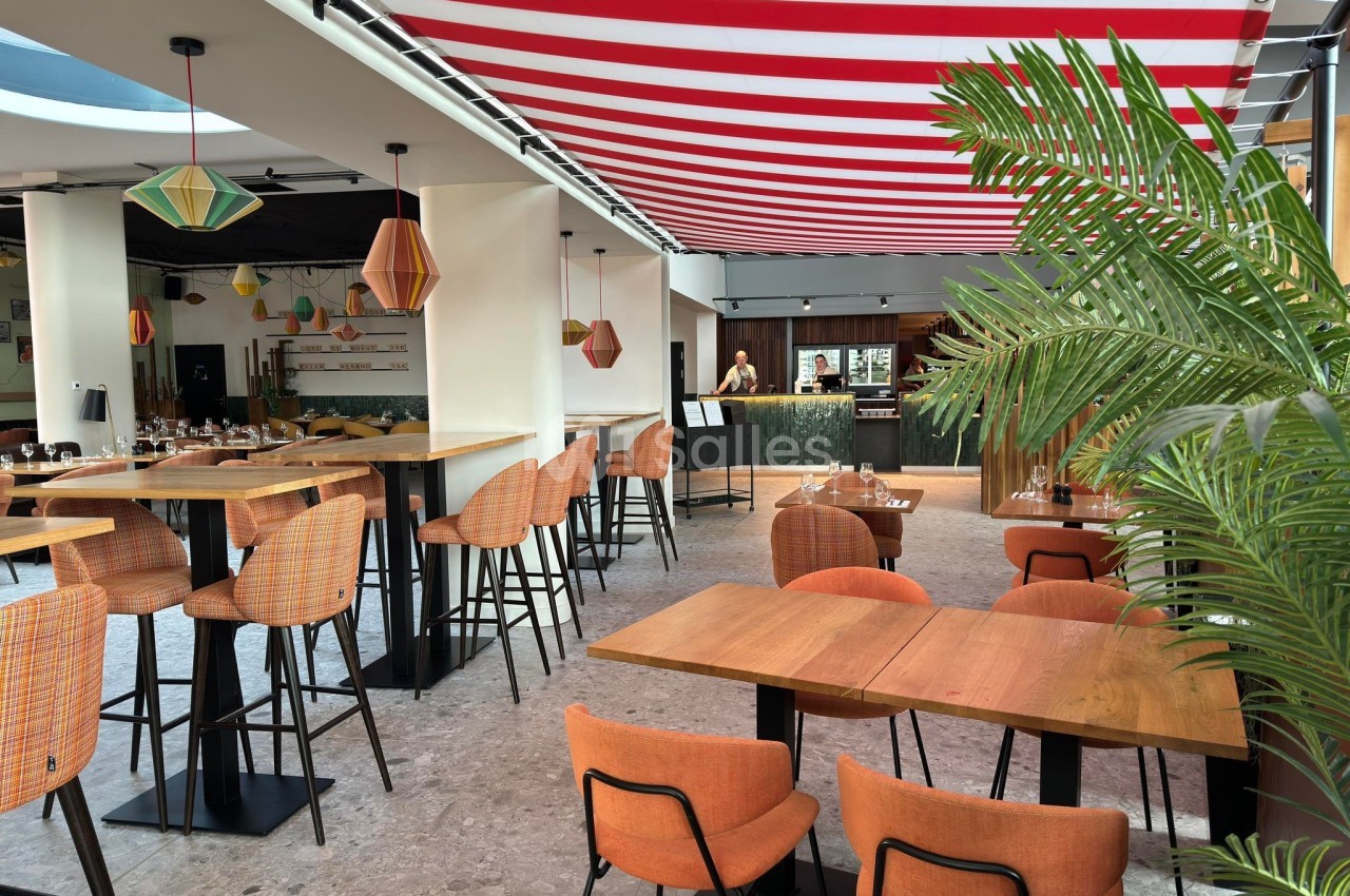 Salle de restaurant lumineuse avec tables en bois, chaises orange, plantes et un plafond rayé rouge et blanc.