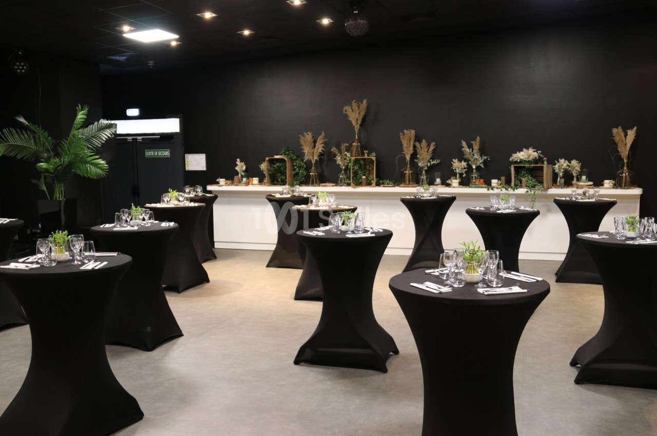 Salle événementielle avec des tables hautes noires dressées, décorations végétales et éclairage tamisé.