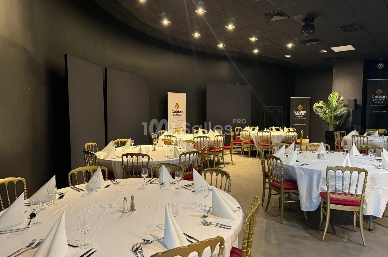 Salle de réception avec tables rondes dressées de nappes blanches, chaises dorées et éclairage tamisé.