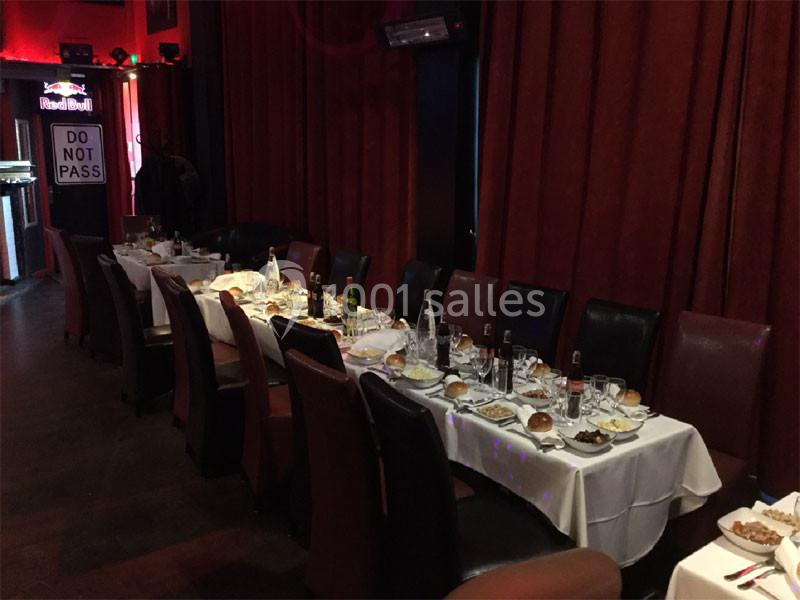 Tables dressées avec nappes blanches, assiettes, verres et couverts, alignées dans une salle aux rideaux rouges.