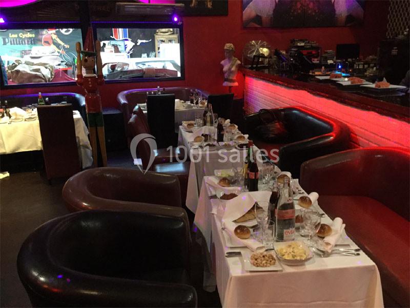 Salle de restaurant avec tables dressées, nappes blanches, bouteilles de vin et décorations lumineuses rouges.