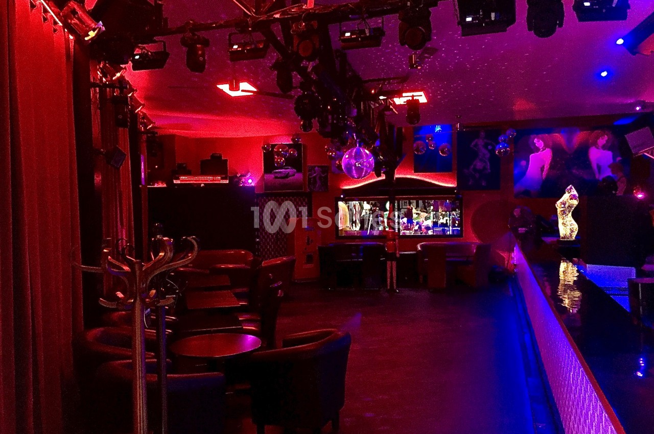 Salle de bar ou discothèque avec éclairage rouge et bleu, tables, fauteuils et bar en arrière-plan.