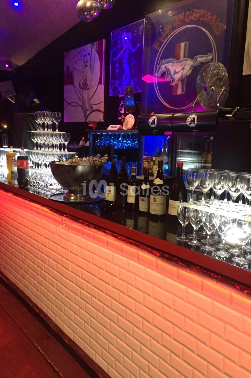 Bar illuminé avec des verres à vin alignés, des bouteilles et une fontaine à champagne sur le comptoir.