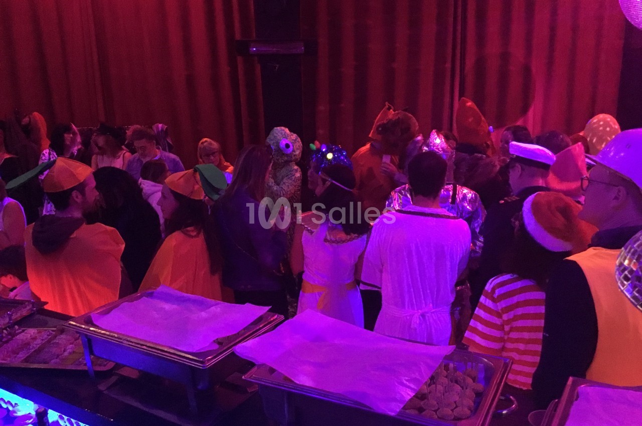 Personnes déguisées rassemblées dans une salle sombre avec des buffets éclairés par une lumière violette.
