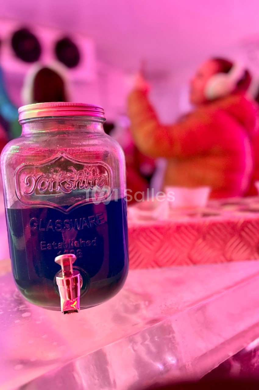 Bocal en verre rempli de liquide bleu avec un robinet, posé sur une surface glacée dans un environnement lumineux rose.