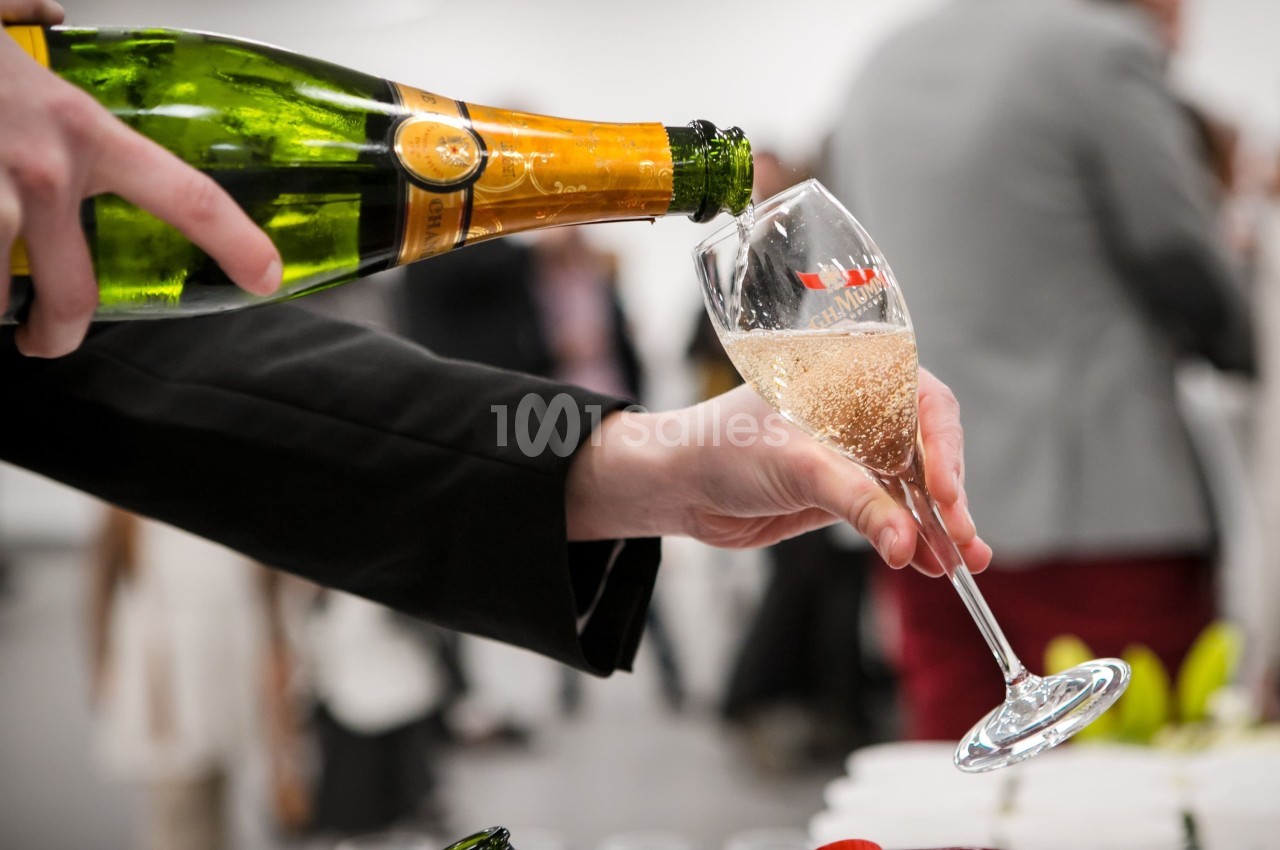 Une main verse du champagne dans une flûte lors d'un événement, avec un arrière-plan flou.